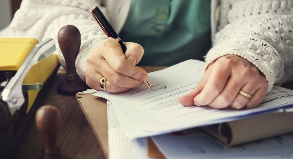 Person signing legal documents with a pen, surrounded by papers and a stapler, illustrating the process of estate planning and legal authorization in a law office setting.