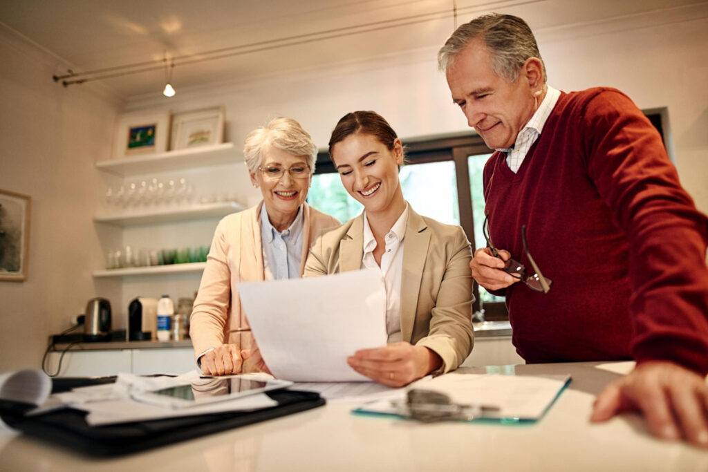 Woman in business attire discussing estate planning documents with an elderly couple, smiling and reviewing papers in a bright, modern kitchen setting.