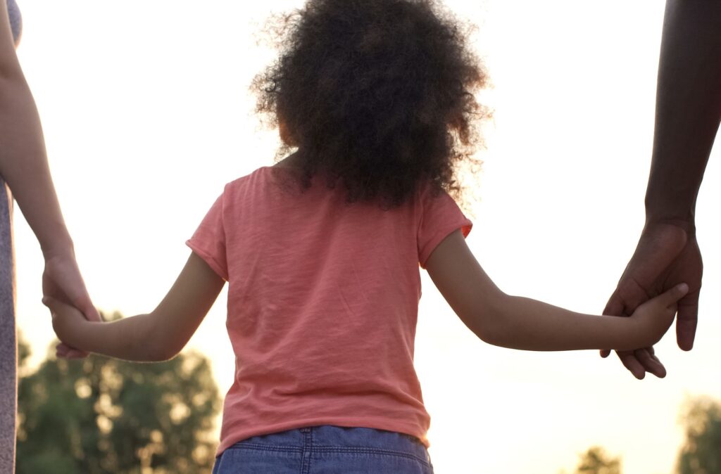 Child holding hands with adults, symbolizing family and adoption, against a sunset backdrop.