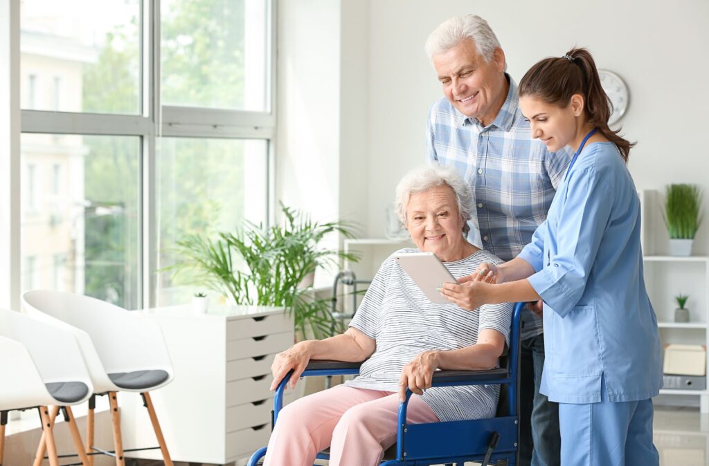 Smiling elderly woman in wheelchair with caregiver and family member, reviewing information on tablet in bright, modern nursing home setting, emphasizing elder care and support.