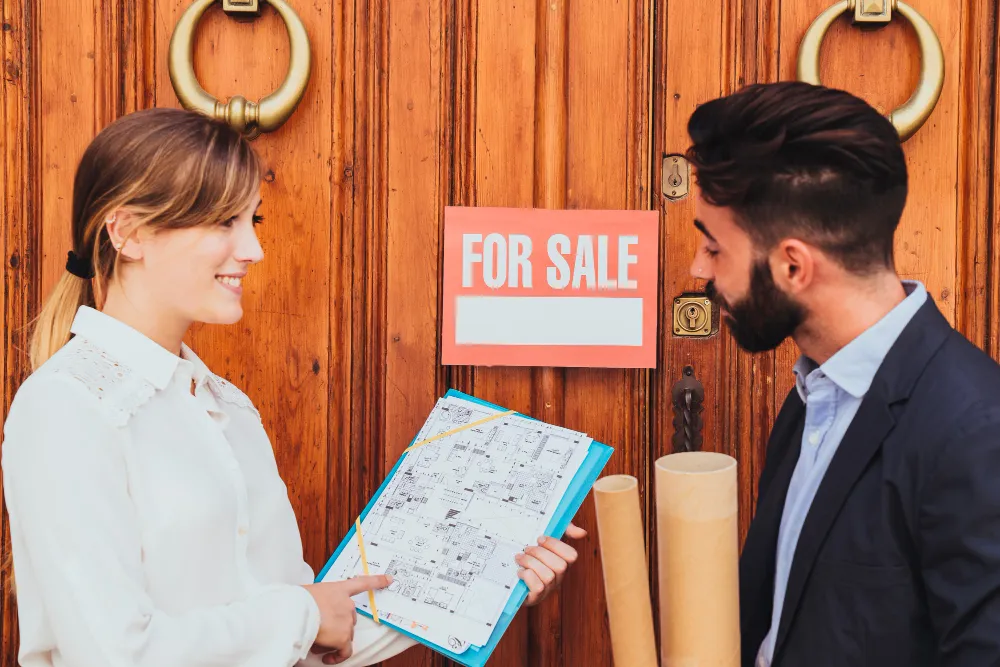 Real estate agents discussing property details in front of a "For Sale" sign, with architectural plans and scrolls in hand, emphasizing property sales and legal considerations in Oklahoma.