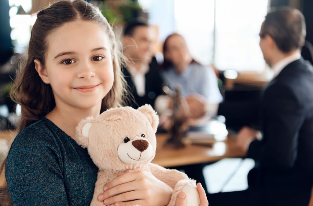 Smiling girl holding a teddy bear in a law office, representing the emotional aspect of guardianship and care for children in legal contexts.